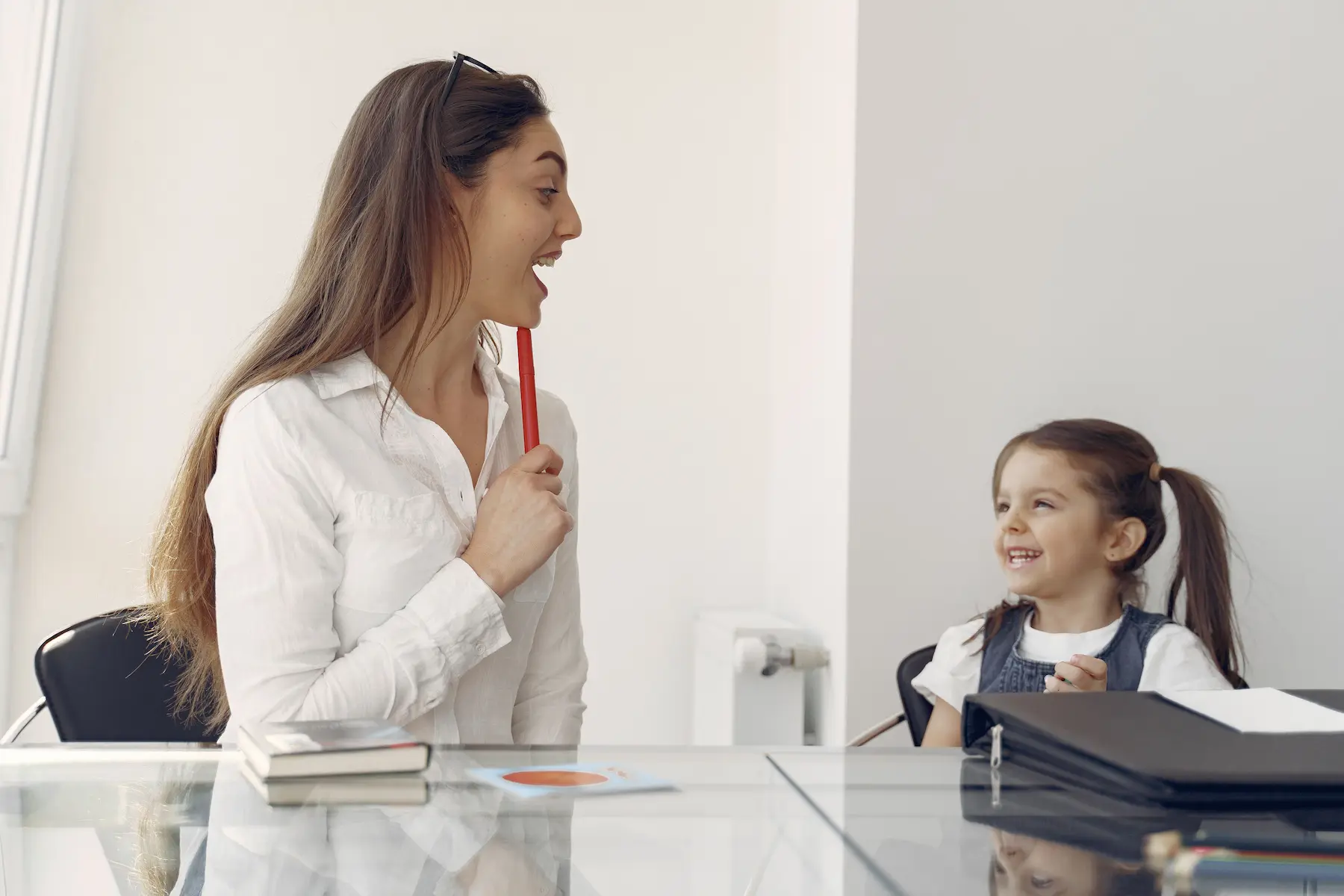 Adult woman on the left and a girl on the right seated at a table. The woman is pointing to her chin to show the girl how to position her mouth during a speech therapy session for childhood apraxia of speech (CAS). The girl is smiling up at her.