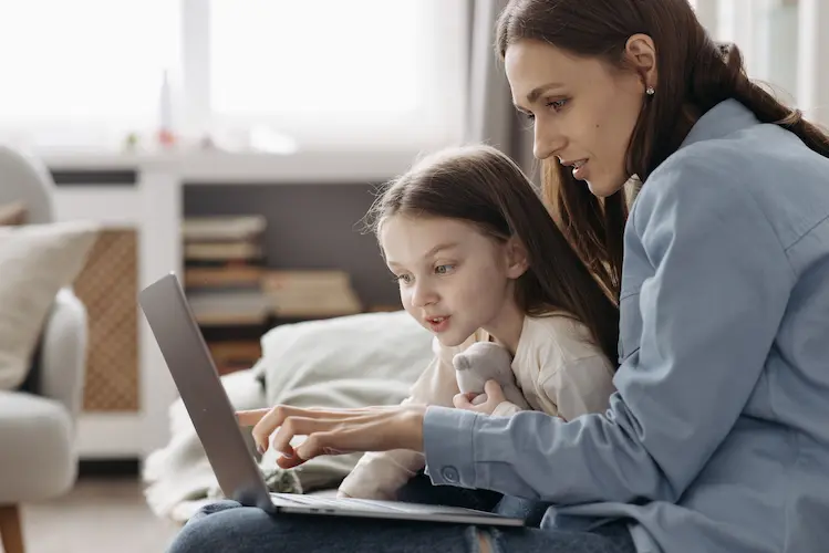 Image of speech therapy services for a mother and young daughter sitting on the sofa with a laptop. They are staring at the screen as part of their online speech therapy session by telehealth.