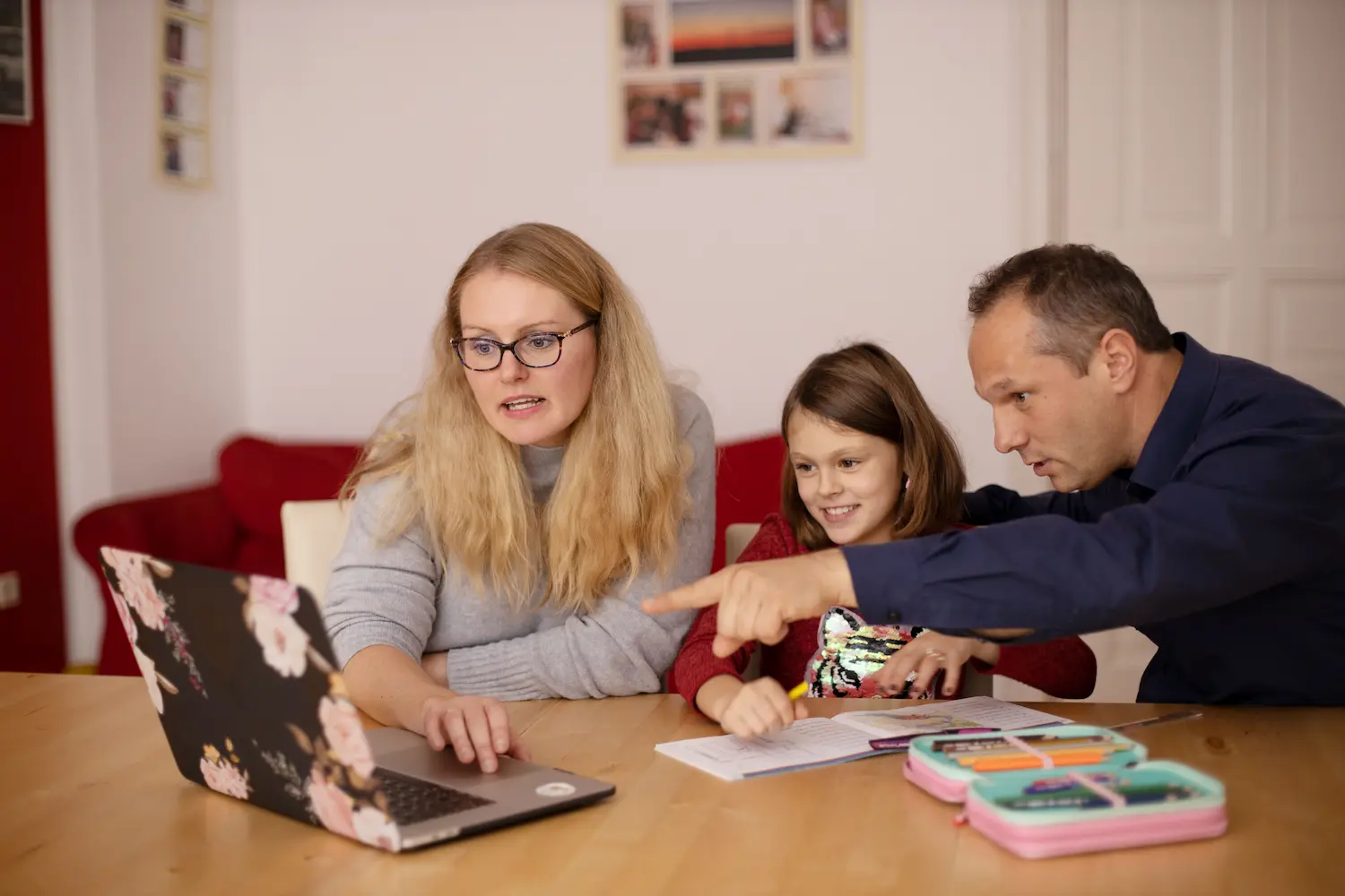 Mother and father with their young daughter at the table. They are viewing a laptop and the father is pointing at the screen as part of their childhood apraxia of speech (CAS) speech therapy session by telehealth.